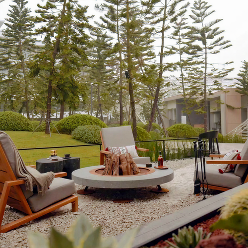 Outdoor patio seating arrangement with three wooden lounge chairs and a round concrete fire pit table on a gravel surface, surrounded by tall pine trees and greenery.