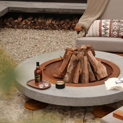 A round concrete fire pit table with a wood-burning fire and a built-in burner, styled in an outdoor patio setting with a bottle, glasses, and a candle on a tray.